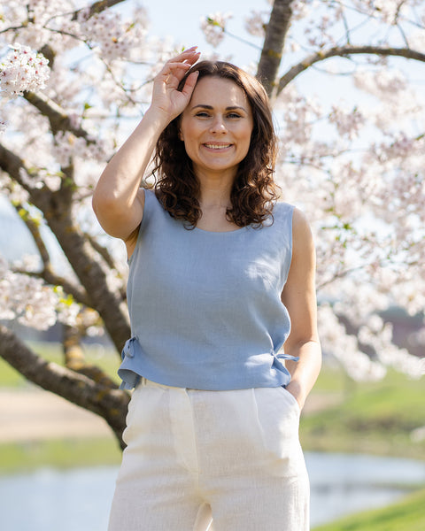 Una mujer sonriente con cabello oscuro y ondulado lleva una blusa sin mangas de lino azul nublado con lazos laterales, combinada con pantalones blancos de lino. Está al aire libre bajo un cerezo en flor, con un cuerpo de agua y un paisaje verde en el fondo suave.