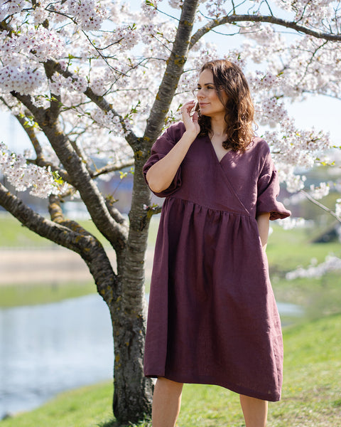 Una mujer sonriente con cabello oscuro y ondulado está al aire libre junto a un cerezo en flor. Lleva un vestido cruzado de lino color púrpura sombra hasta la rodilla, con mangas hasta el codo. Su mano derecha está levantada hacia su rostro. Al fondo, hay un cuerpo de agua y césped verde bajo un cielo despejado.