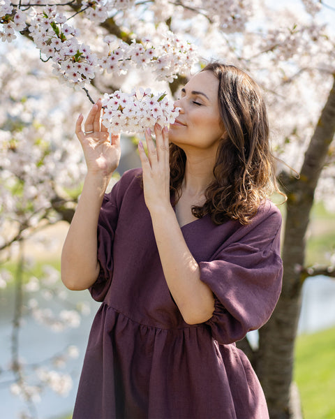 Una mujer sonriente con cabello oscuro y ondulado aparece en un primer plano al aire libre, acercando suavemente un racimo de flores blancas de cerezo a su nariz para olerlas. Tiene los ojos cerrados y parece disfrutar de la fragancia. Lleva un vestido de lino color púrpura sombra con escote en V y mangas abullonadas hasta el codo. De fondo se ven más flores de cerezo y un cuerpo de agua.
