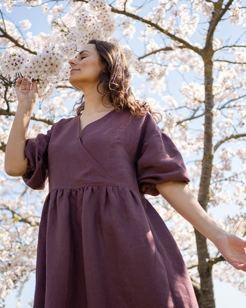 Una persona con cabello oscuro y ondulado, con los ojos cerrados, está al aire libre, oliendo las flores blancas de un árbol. Lleva un vestido largo de cuello en V color púrpura sombra, con mangas sueltas hasta el codo. Las ramas del árbol en flor se ven detrás de ella contra un cielo azul brillante.