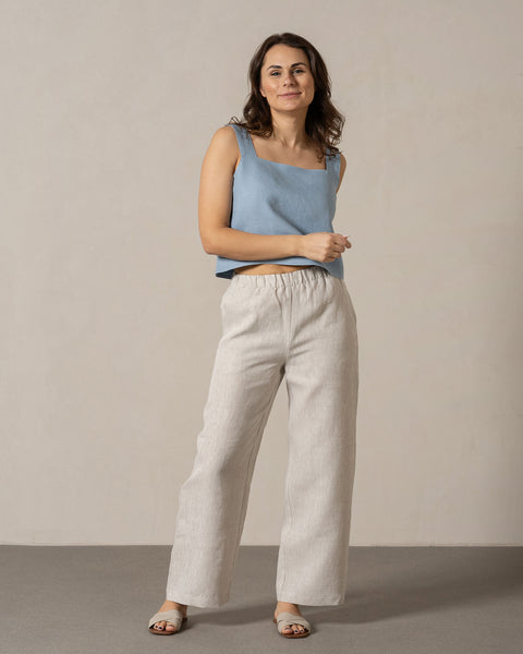 Una mujer de piel de tono medio y cabello oscuro y ondulado está de pie frente a la cámara con una leve sonrisa. Lleva una blusa sin mangas de lino menique con escote cuadrado y pantalones anchos de lino con cintura elástica. Sus manos descansan casualmente a los lados y lleva sandalias de color claro. Está sobre una superficie gris claro frente a una pared lisa de color beige claro.