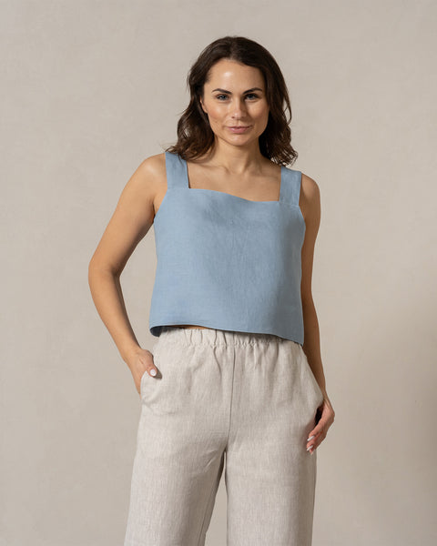 Una mujer de piel de tono medio y cabello oscuro y ondulado mira directamente a la cámara con una expresión amable. Lleva una blusa sin mangas de color azul nublado con escote cuadrado y pantalones anchos de color natural con cintura elástica, y sus manos están colocadas de manera casual en los bolsillos. El fondo es de un color beige claro y liso.