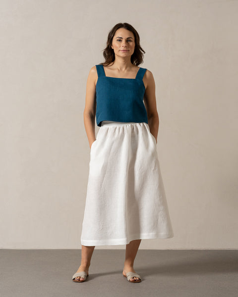 Una mujer de piel tono medio y cabello oscuro hasta los hombros está de pie mirando hacia adelante contra una pared lisa de color claro. Lleva una blusa sin mangas color verde azulado con escote cuadrado y una falda blanca de Menique, con las manos en los bolsillos. Está descalza y parece estar sobre un suelo gris claro.