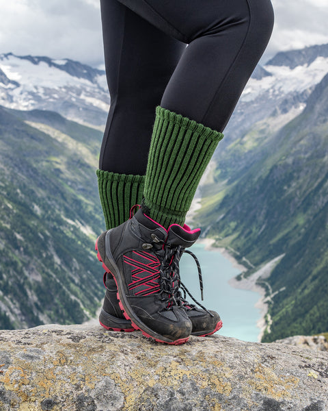 Primer plano de un senderista que lleva calcetines de senderismo de merino verde acanalados con botas de senderismo negras y rosas, de pie sobre una roca con vistas a un lago alpino y montañas.