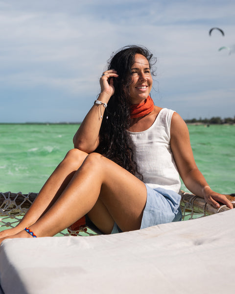 Una mujer se sienta al sol, vistiendo una camiseta sin mangas blanca, pantalones cortos azules y una bufanda naranja, con un fondo de cielo brillante y agua turquesa.