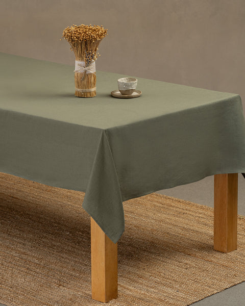 El mantel de lino verde piedra de menique sobre una mesa de madera. Encima del mantel hay un jarrón hecho de pajillas naturales y una taza de barro. Debajo de la mesa hay una alfombra.
