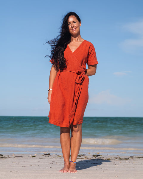 A woman with long dark hair wearing a cinnamon red linen wrap dress stands barefoot on a sandy beach, looking slightly to her right with a calm expression. The ocean with gentle waves is in the background under a bright, partly cloudy sky.