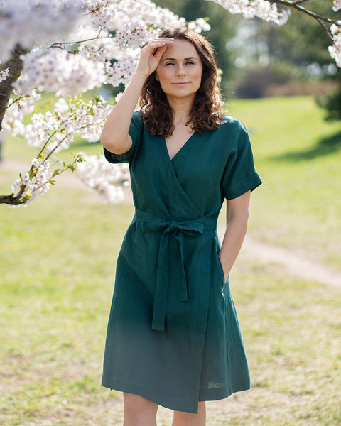 Una mujer de cabello castaño, vestida con un vestido cruzado de manga corta de color verde oscuro, está de pie al aire libre bajo un árbol con flores blancas.