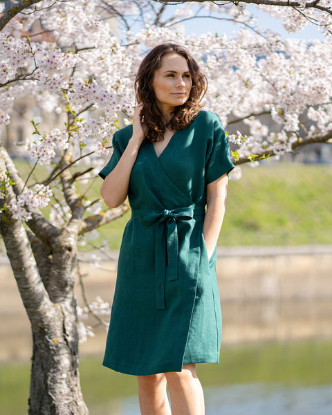 Una mujer de cabello castaño, vestida con un vestido cruzado de manga corta de color verde oscuro, está de pie al aire libre bajo un árbol con flores blancas.