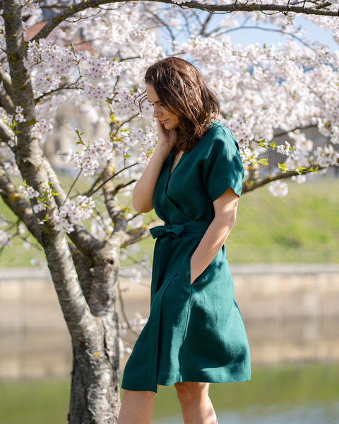 Una mujer de cabello castaño, vestida con un vestido cruzado de manga corta de color verde oscuro, está de pie al aire libre bajo un árbol con flores blancas.