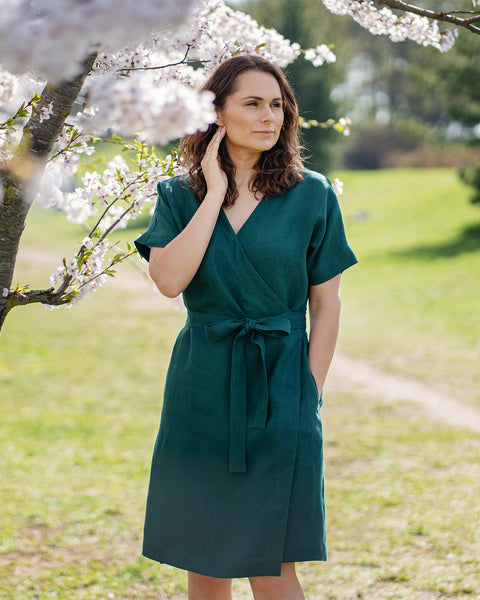 Una mujer de cabello castaño, vestida con un vestido cruzado de manga corta de color verde oscuro, está de pie al aire libre bajo un árbol con flores blancas.