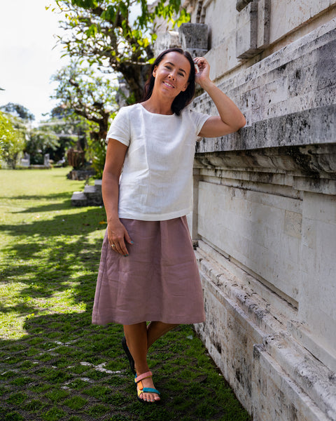 Una mujer de cabello oscuro sonríe mientras se apoya en una pared de piedra blanca al aire libre. Lleva una blusa blanca de manga corta, una falda rosa empolvado hasta la rodilla y sandalias planas de colores. Su codo izquierdo descansa sobre la pared y su mano izquierda está cerca de su cabello. Al fondo se ve una zona de césped y árboles bajo un cielo soleado.
