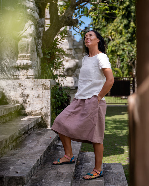 Una mujer de cabello oscuro está de pie en unos escalones de piedra al aire libre, mirando hacia arriba. Lleva una blusa blanca de manga corta, una falda rosa empolvado hasta la rodilla y sandalias planas de colores. Tiene las manos en los bolsillos de la falda. A su lado hay un muro de piedra desgastado con grabados, y al fondo se ven árboles con hojas verdes bajo un cielo despejado.