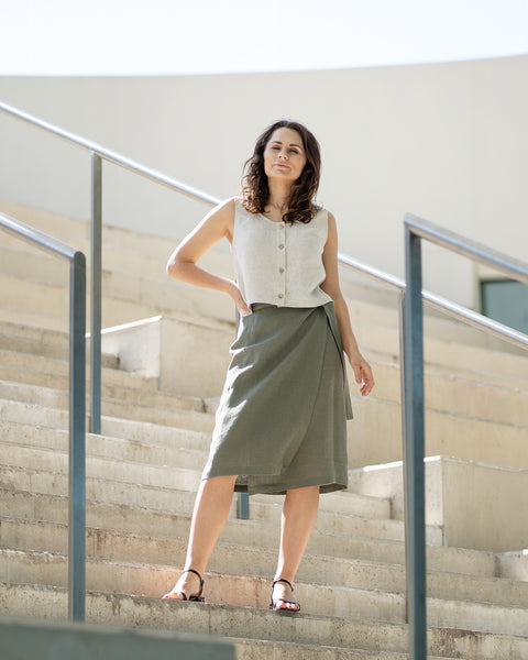 Una mujer de cabello castaño, vestida con una blusa sin mangas de lino natural con botones al frente y una falda cruzada verde piedra, posa en unas escaleras de concreto al aire libre.