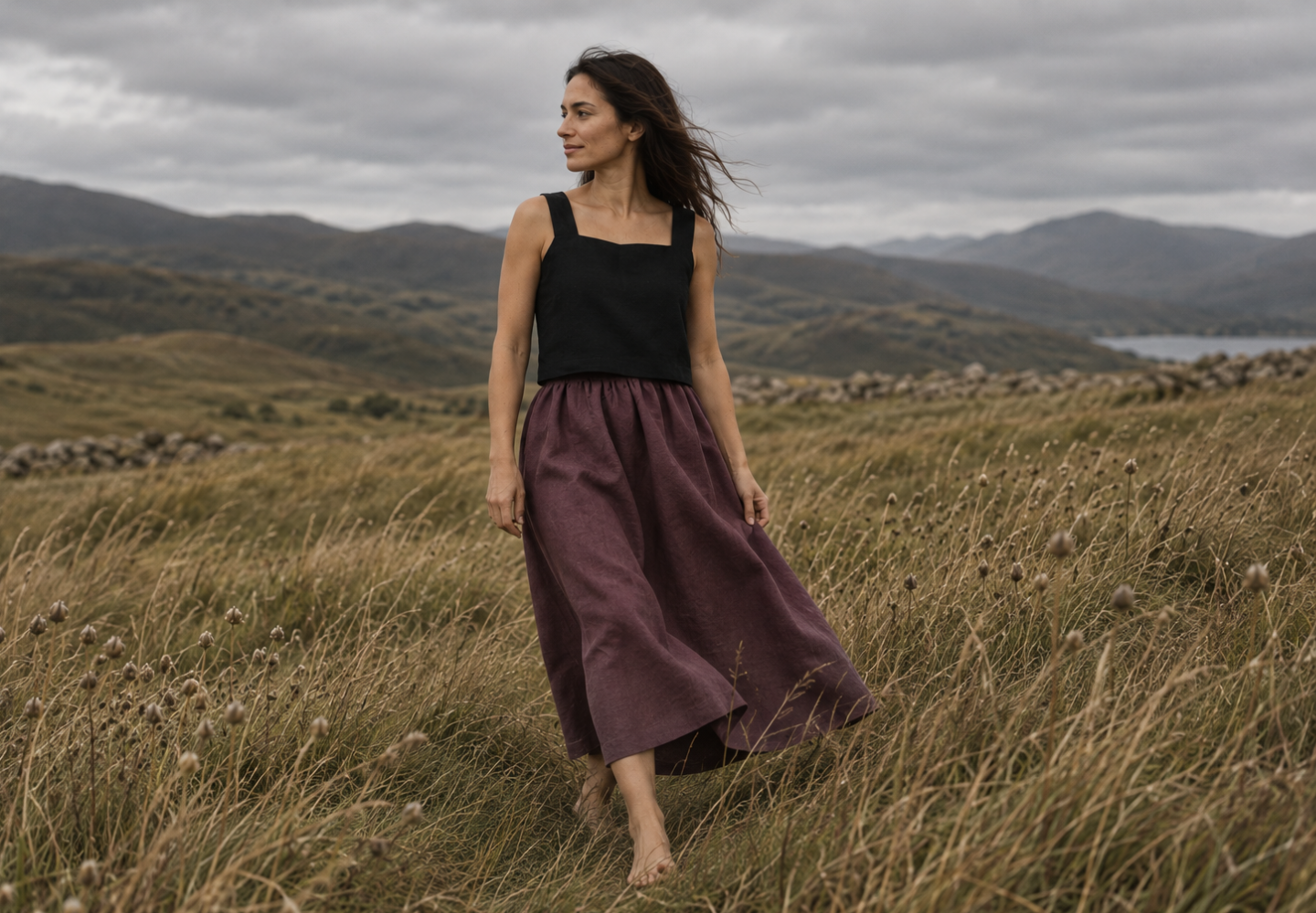 A woman walking barefoot through a field of tall grass, wearing a black square-neck top and a long plum-colored linen skirt, with a hilly landscape behind her.