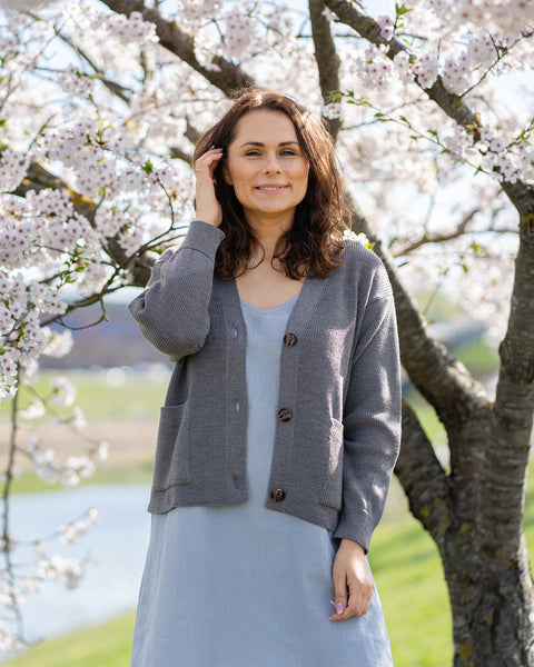 Una mujer de cabello castaño, que lleva un cárdigan gris de punto sobre un vestido azul claro, está de pie al aire libre junto a un árbol con flores blancas.