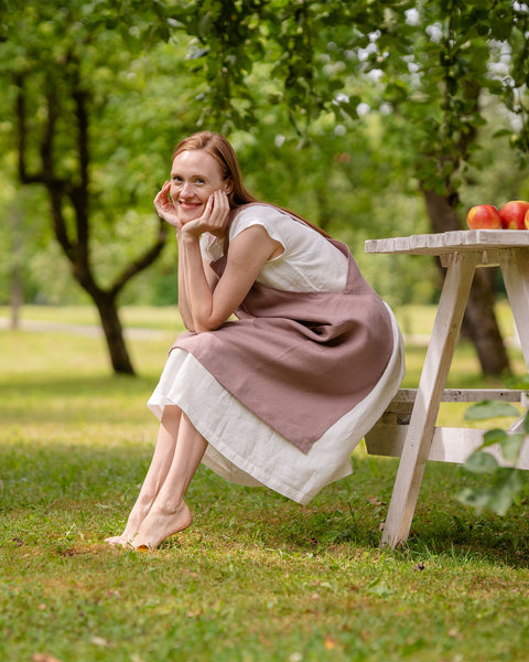 Una mujer de piel clara con cabello largo rojizo que lleva un delantal de lino menique está sentada descalza en un banco blanco en un jardín soleado, sonriendo con la barbilla apoyada en las manos. A su lado hay una pequeña mesa con manzanas y flores.