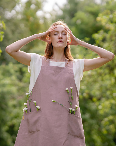 Una persona de piel clara con cabello rojizo está al aire libre, vistiendo un delantal de lino color rosa desvaído con flores blancas en los bolsillos, con las manos tocándose la cabeza, frente a un fondo de vegetación desenfocada.