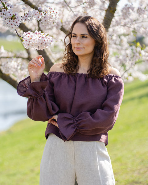 Una mujer de cabello castaño está al aire libre bajo un cerezo en flor, mirando hacia su derecha y sonriendo. Lleva una blusa con los hombros descubiertos y mangas largas y sueltas recogidas en las muñecas, y pantalones anchos de color claro. El fondo es un suave desenfoque de césped verde y más cerezos en flor.
