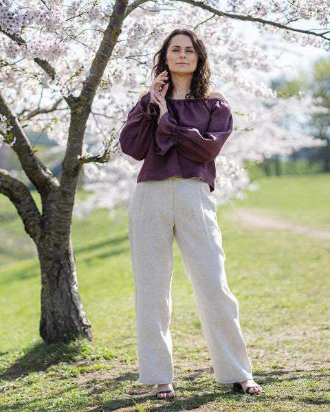 Una mujer de piel clara y cabello oscuro y ondulado, que lleva una blusa morado sombra, con los hombros descubiertos, se muestra desde la cintura hacia arriba.