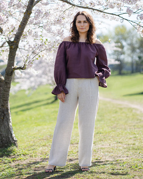 Una mujer de piel clara y cabello oscuro y ondulado, que lleva una blusa morado sombra, con los hombros descubiertos, se muestra desde la cintura hacia arriba. Ella mira directamente al espectador con una leve sonrisa.