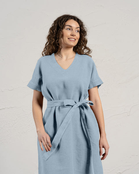 Una mujer con cabello rizado y castaño sonríe mientras modela un vestido menique de lino azul nublado con mangas cortas y bolsillos.
