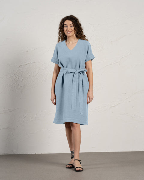 Una mujer con cabello rizado y castaño sonríe mientras modela un vestido menique de lino azul nublado con mangas cortas y bolsillos.