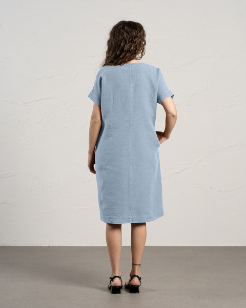 La vista trasera de una mujer con cabello rizado y castaño muestra que lleva un vestido de lino azul nublado con mangas cortas y bolsillos, de pie contra una pared texturizada de color blanco roto.