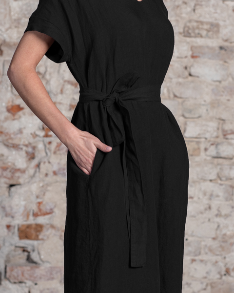 Una mujer luce un vestido suelto de lino negro puro de menique con cintura anudada y una mano en el bolsillo lateral, de pie frente a una pared de ladrillo texturizada.