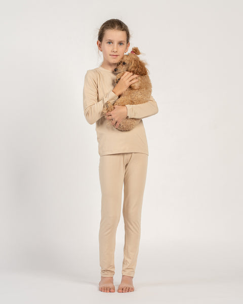 Niña con una camiseta de manga larga y pantalones beige de bambú, de pie descalza y sosteniendo en sus brazos un pequeño caniche marrón.