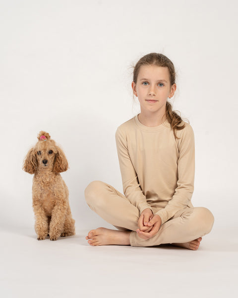 Niña sentada con las piernas cruzadas, con camiseta y pantalones largos de bambú beige, junto a un pequeño caniche marrón con una coleta rosa.