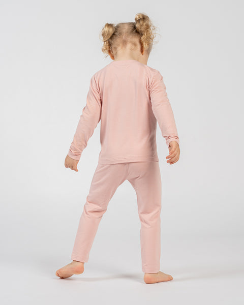 Niño con camiseta de manga larga rosa y pantalones a juego, de pie descalzo con la espalda hacia la cámara y los brazos relajados a los lados, mostrando el conjunto desde atrás sobre un fondo liso.