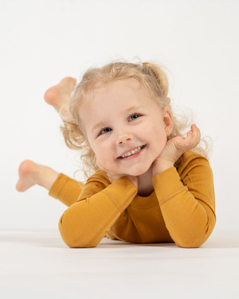 A smiling child with curly blonde pigtails, dressed in a yellow long-sleeved top and matching pants, is lying on their stomach on a white surface. The child's head is propped up by their hands, and they are looking towards the camera with a joyful expression. Their bare feet are raised slightly in the background.