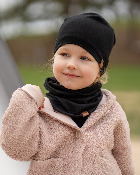 Un niño pequeño de piel clara y cabello rubio mira ligeramente hacia la derecha con una sonrisa suave, llevando un gorro negro de menique y una braga de cuello negra a juego. Viste un abrigo de textura acogedora en tono rosa claro o beige, con un fondo exterior desenfocado.