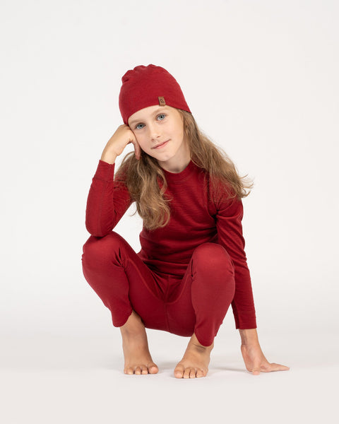 una niña, agachada, con un gorro de merino para niños en color cereza real, y un conjunto a juego de merino con camiseta de manga larga y pantalones.