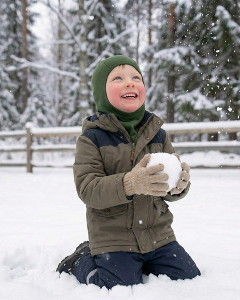 Niño sonriente vestido con ropa de abrigo de invierno y una braga para la cabeza de lana merina verde, jugando con una bola de nieve en un bosque nevado con copos de nieve cayendo.