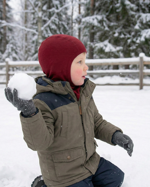 Niño pequeño con una chaqueta de invierno cálida y una braga para la cabeza de lana merina color burdeos, sosteniendo una gran bola de nieve mientras está de rodillas en la nieve fresca en un paisaje invernal boscoso.