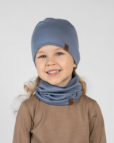 A studio portrait of a smiling young blonde girl looking directly at the camera showing her teeth. She is wearing a matching set of a dusty blue beanie hat and a dusty blue neck gaiter gathered comfortably around her neck. Both items feature a small brown leather logo patch. She is wearing a light brown long-sleeved shirt against a plain light grey background.