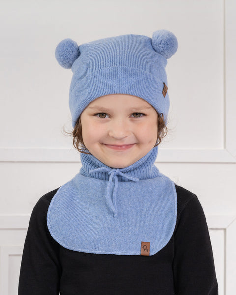 Un niño pequeño con cabello rizado castaño claro y una sonrisa sutil, mira directamente al espectador. Lleva un gorro de punto azul claro con dos pompones azul claro en la parte superior y una pequeña etiqueta marrón en un lado, y una bufanda de punto azul claro a juego con un lazo, sobre una camiseta negra de manga larga. El fondo es una pared blanca con paneles.