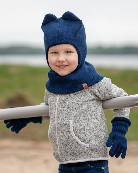 Un niño pequeño de piel clara y cabello claro sonríe a la cámara, apoyado en una barra horizontal de color claro al aire libre. Lleva una chaqueta gris jaspeada con cremallera, vaqueros azules y accesorios menique tejidos en azul oscuro, que incluyen un gorro estilo pasamontañas con dos orejas redondeadas en la parte superior y un par de guantes acanalados. En el fondo desenfocado, se ve un cuerpo de agua y una orilla verde tenue.