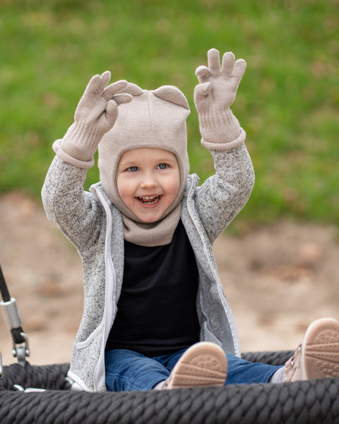 Un niño alegre, con ojos azules y una sonrisa abierta, está sentado en un columpio de cuerda negra con los brazos levantados. Lleva una braga de cuello menique color beige cremoso con orejas, una bufanda a juego, guantes beige, una sudadera con cremallera gris jaspeada, vaqueros azules y zapatillas rosas. El fondo es césped verde desenfocado y tierra marrón.