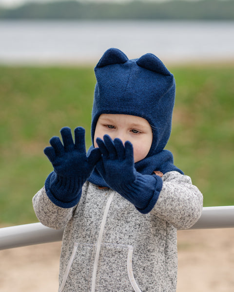 Un niño pequeño que lleva una braga para la cabeza menique azul oscuro con orejas, guantes azul oscuro y una sudadera con cremallera gris jaspeada, sostiene las manos frente a su rostro, mirando ligeramente hacia abajo. El fondo es una escena al aire libre desenfocada con césped verde y un cuerpo de agua.