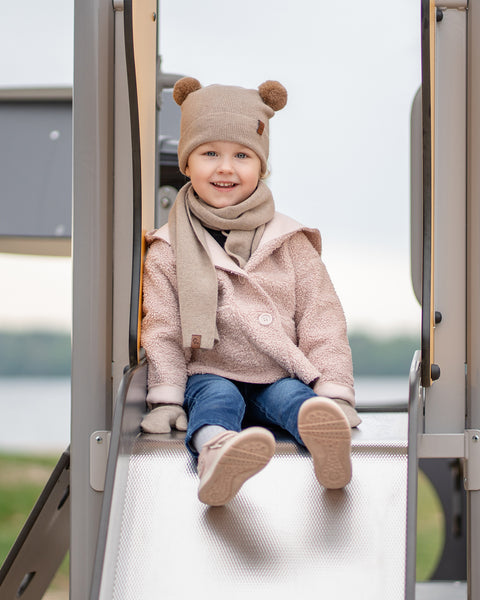 Un niño pequeño con una sonrisa alegre está sentado en la cima de un tobogán de metal en el parque infantil. Lleva puesta una pasamontañas beige con orejas de pompón marrones, una bufanda beige, un abrigo estilo sherpa de color rosa claro, vaqueros azules y zapatillas rosa claro. El fondo desenfocado muestra agua y árboles.