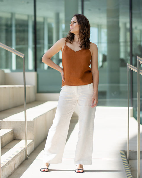 Una mujer de cabello castaño lleva una blusa de tirantes con escote en V color almendra y pantalones blancos, posando en el interior frente a grandes ventanales de cristal.