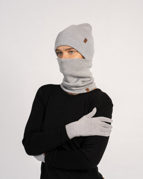 Mujer con un gorro de punto gris claro, guantes a juego y un braga para el cuello que le cubre el rostro, vestida con una camiseta negra de manga larga, posando con los brazos cruzados frente a un fondo liso.