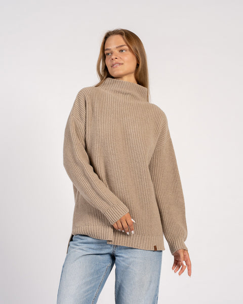 Una mujer posando con un suéter de punto beige de corte holgado y mangas largas, girando ligeramente hacia un lado.