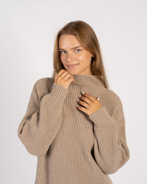 Primer plano de una mujer sosteniendo suavemente el cuello alto de un suéter de punto beige, mostrando la acogedora textura acanalada.