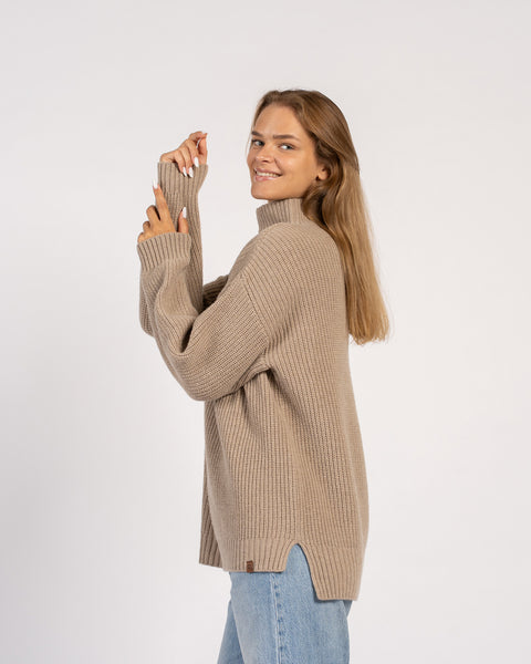 Una mujer sonriente que lleva un suéter de cuello alto de punto suelto en color beige con aberturas laterales, combinado con vaqueros azul claro.