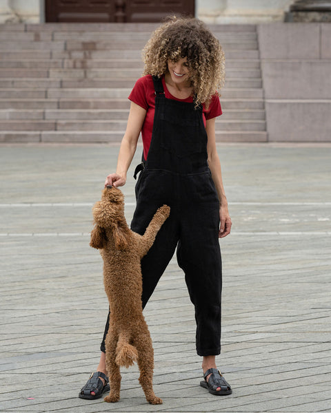 Beautiful woman with curly hair posing in the city wearing pure black linen jumpsuit Nicci, royal cherry merino wool top. Posing with a brown poodle dog.