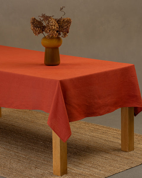 El mantel de lino rojo canela de menique sobre una mesa de madera. Encima del mantel hay un jarrón hecho de pajas naturales y una taza de barro. Debajo de la mesa hay una alfombra.